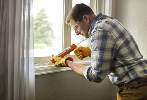 Man in a plaid shirt caulking a window inside a house