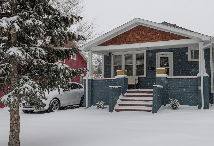 Snow covering the ground view of blue house with a car sitting next to it and a snow covered tree in the foreground