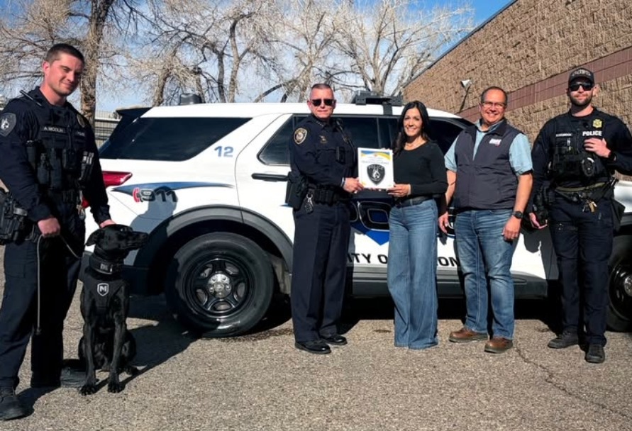 Five people and a dog pose beside a police SUV while presenting an award outdoors.